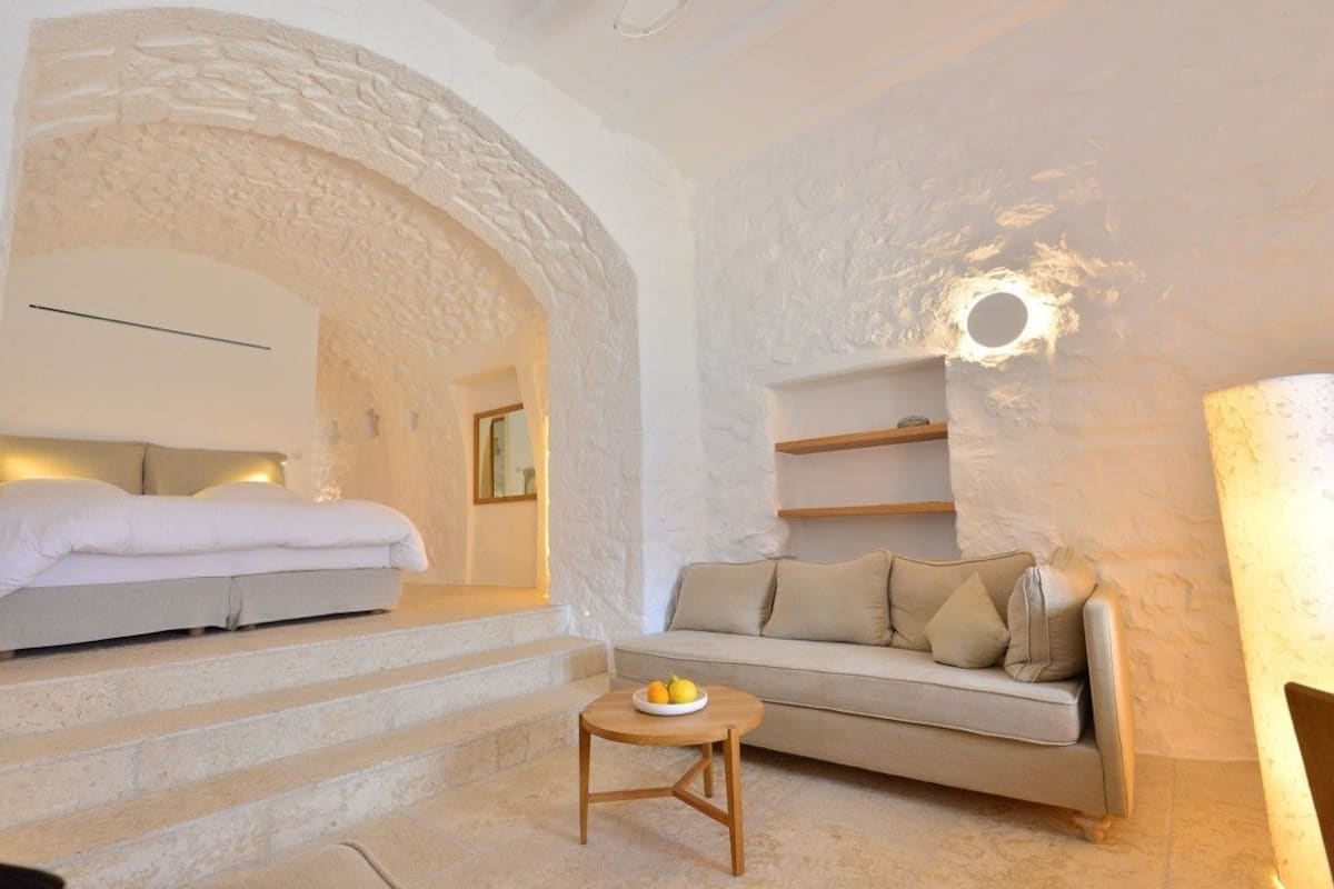Una habitación acogedora con cama, sofá y una mesita baja con fruta. Se aprecia una pared de piedra y una lámpara circular.