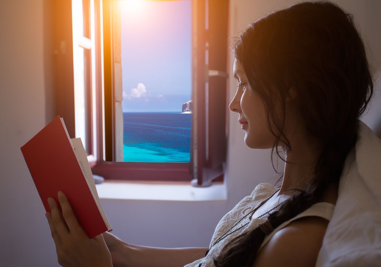 A person reading a book by a window with a view of the ocean.