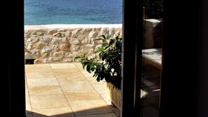 A view from a room with a stone wall and a tiled floor leading to a balcony overlooking a clear blue sea and a rocky hillside. - Pirgos Mavromichali (East Mani)
