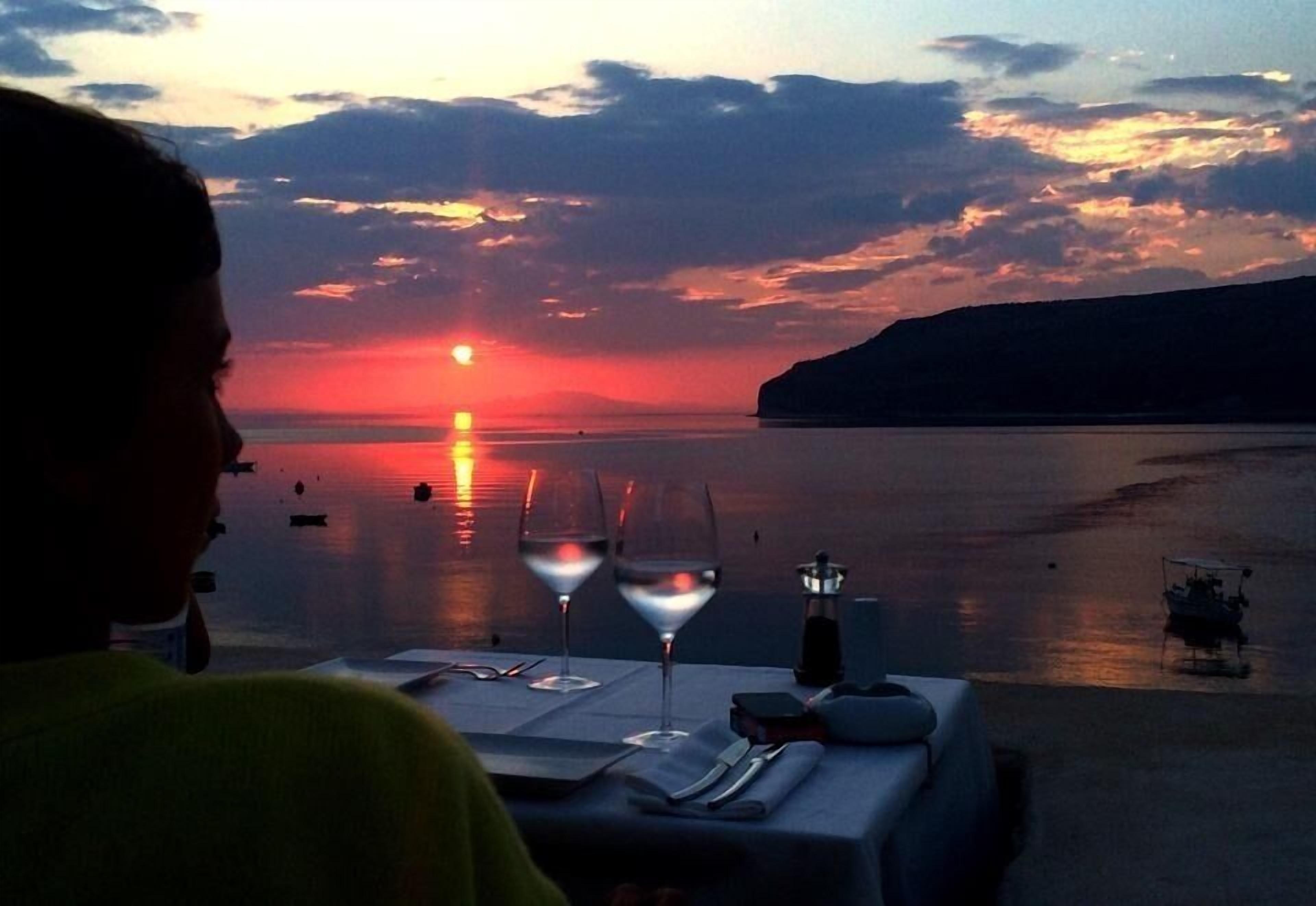 A table set for two with a sunset view, a boat on the water, and a cliff in the background.