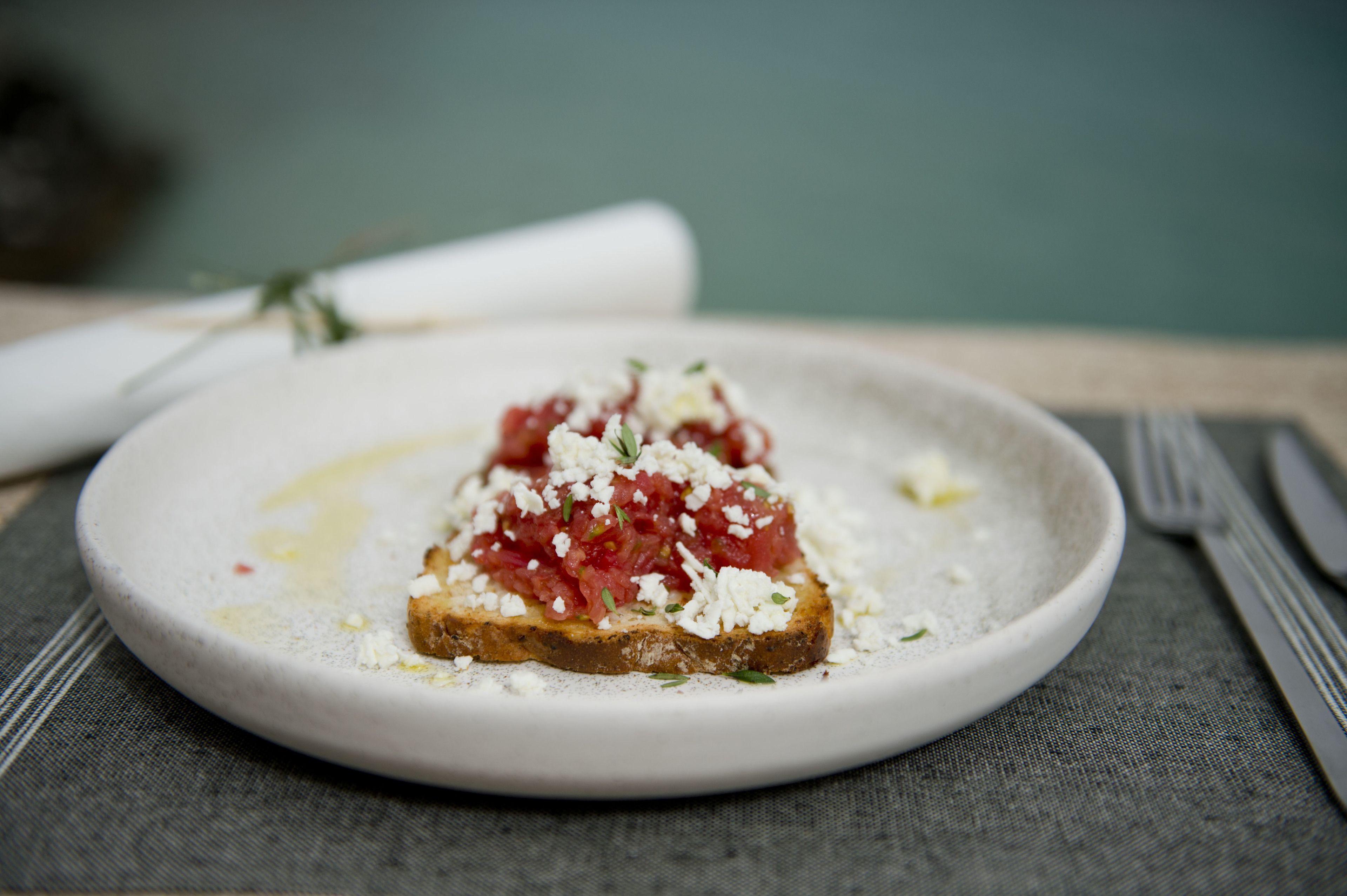 A plate with a slice of toast topped with tomatoes and cheese, accompanied by a fork and knife on a tablecloth.