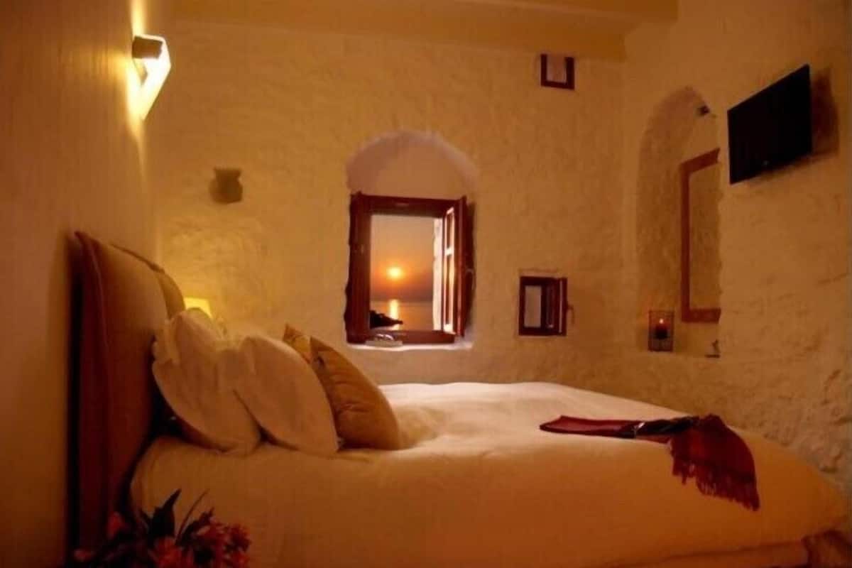 Un dormitorio con cama, almohadas, una mesita de noche, un televisor instalado en la pared y una ventana con vista al atardecer.