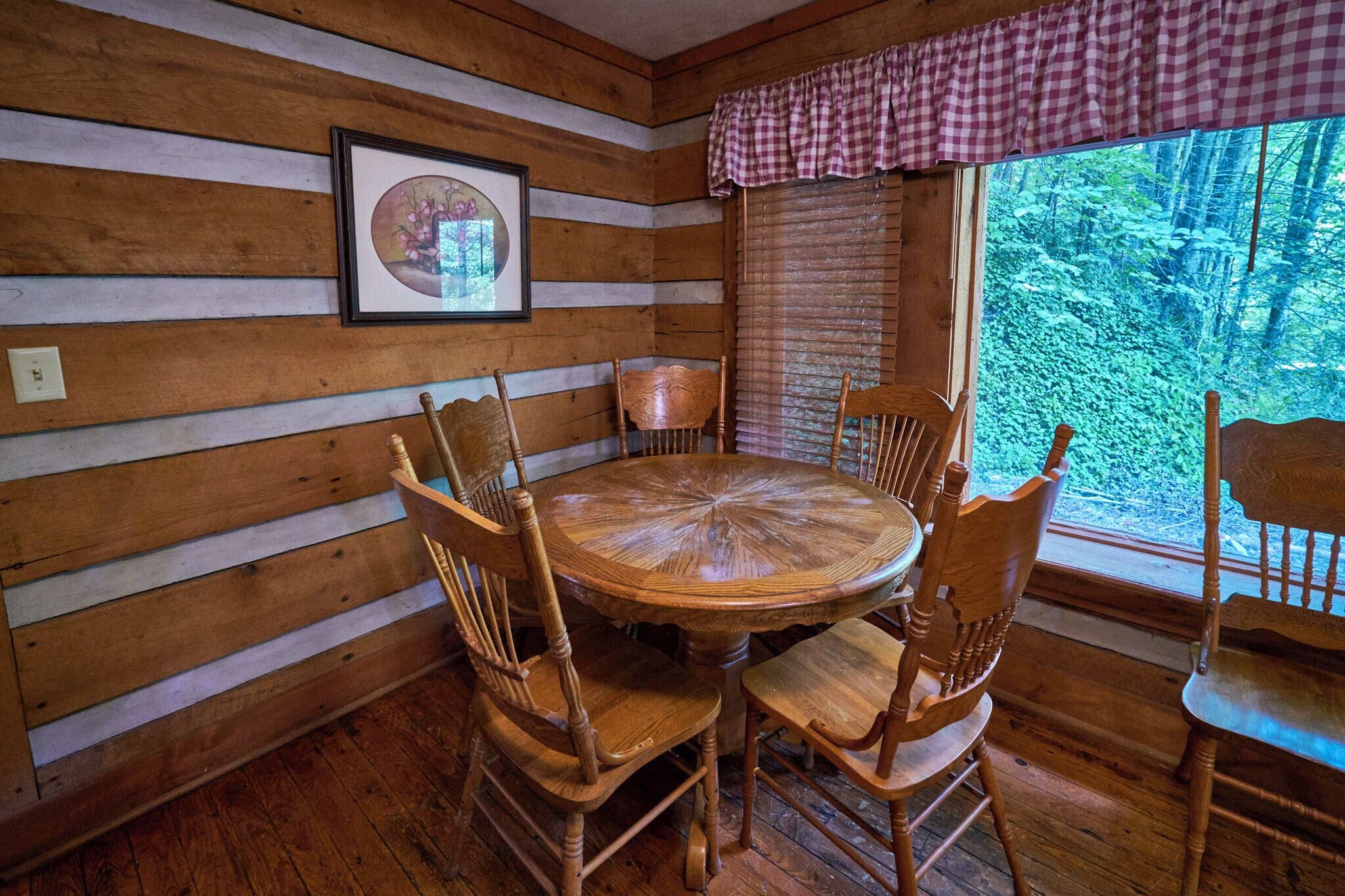 A wooden cabin interior with a round wooden table and six chairs, a framed picture on the wall, and a window with a checkered valance.
