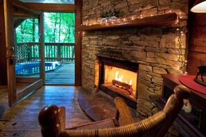 Una habitación acogedora con chimenea de piedra, suelo de madera y vistas a una terraza de madera y árboles.