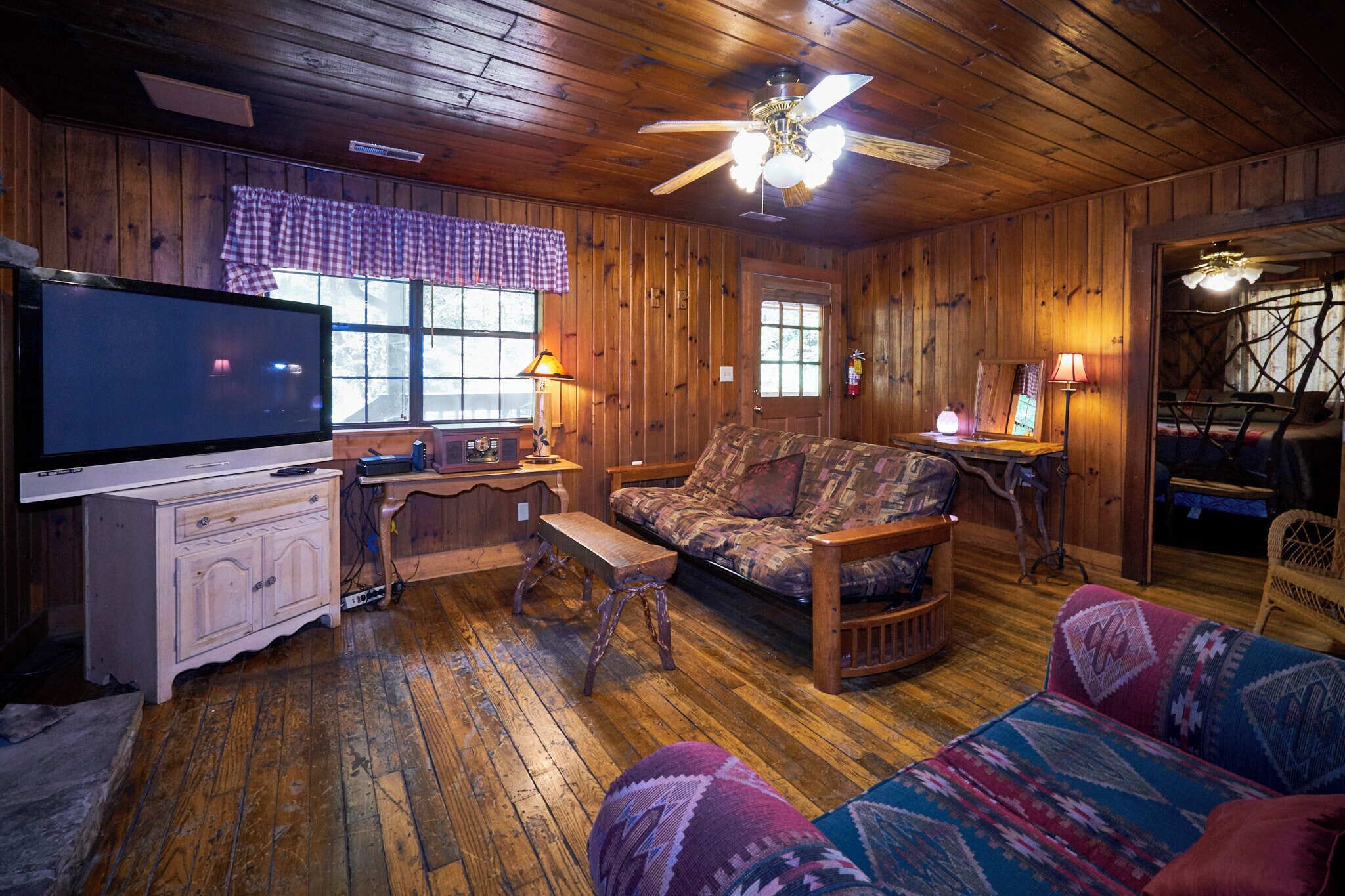 A wooden cabin interior with a television, a sofa, a bench, and a ceiling fan.