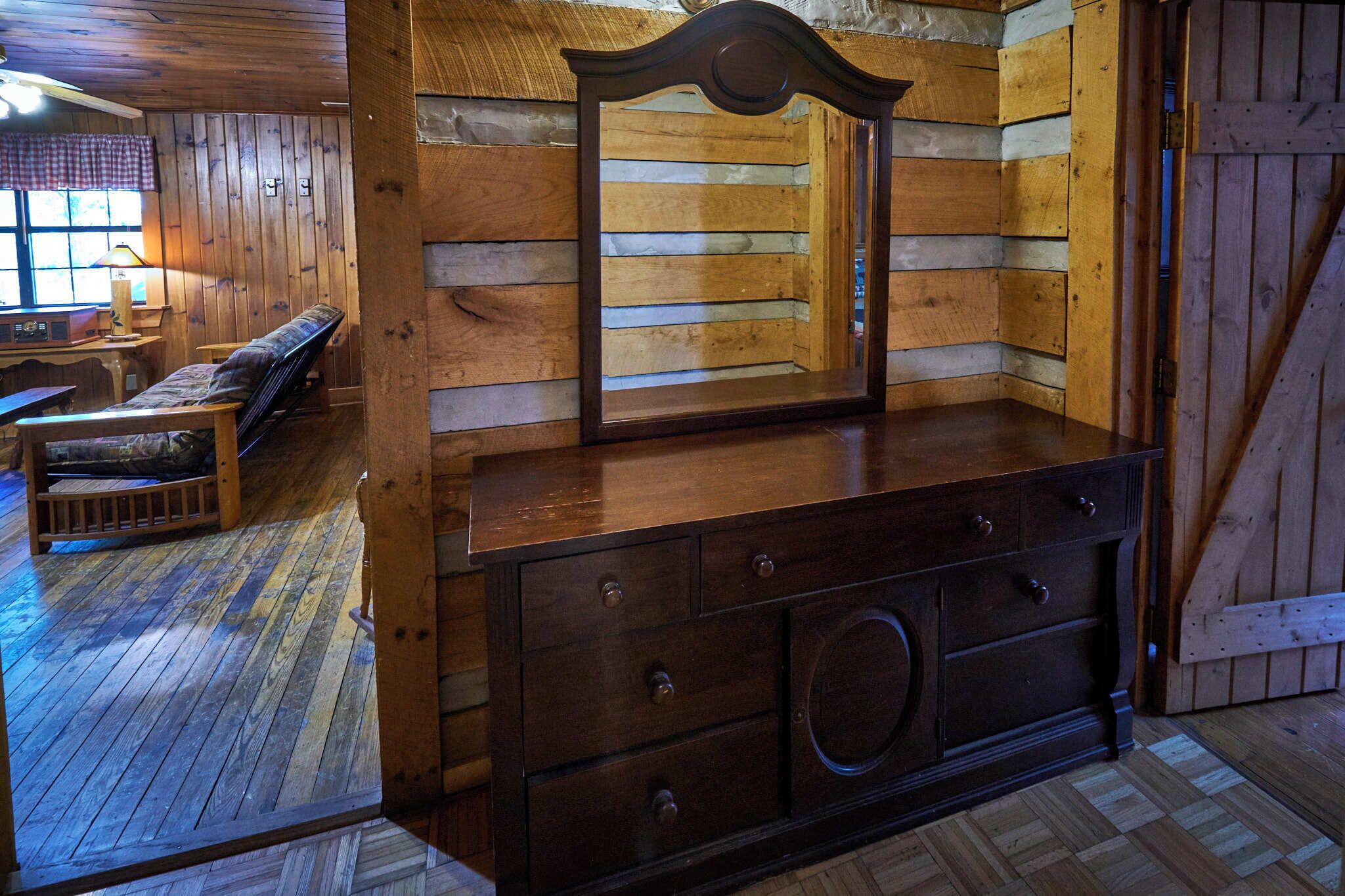 Interior d'una cabana de fusta amb una banca de fusta, un taulell amb mirall i una finestra amb cortina a quadradets.