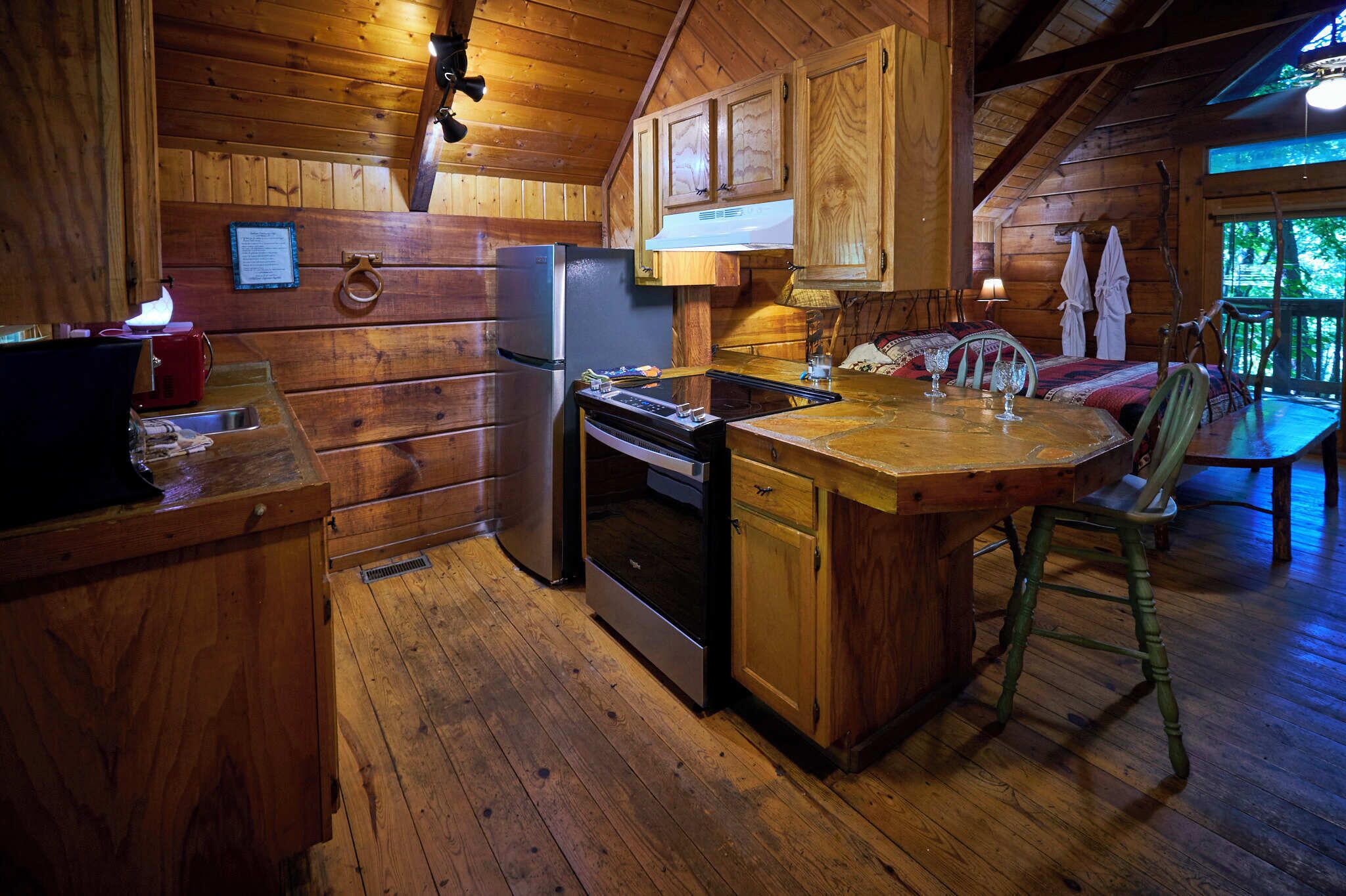 Interno di una baita in legno con cucina, dotata di frigorifero, fornello e lavello. Si nota un tavolo da pranzo con sedie e una finestra vista vegetazione.