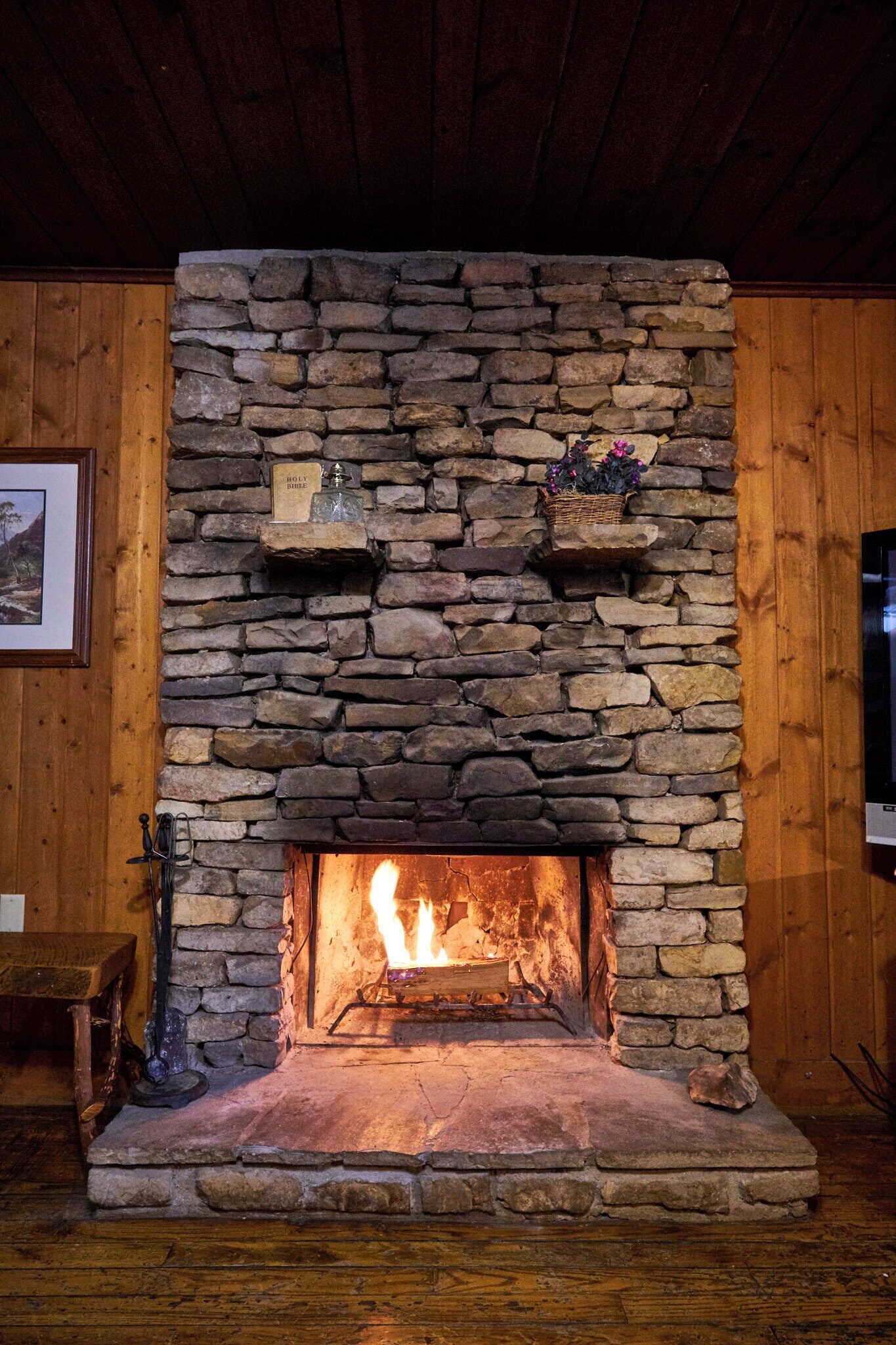 A stone fireplace with a fire, wooden shelves above, and a framed picture on the wall.