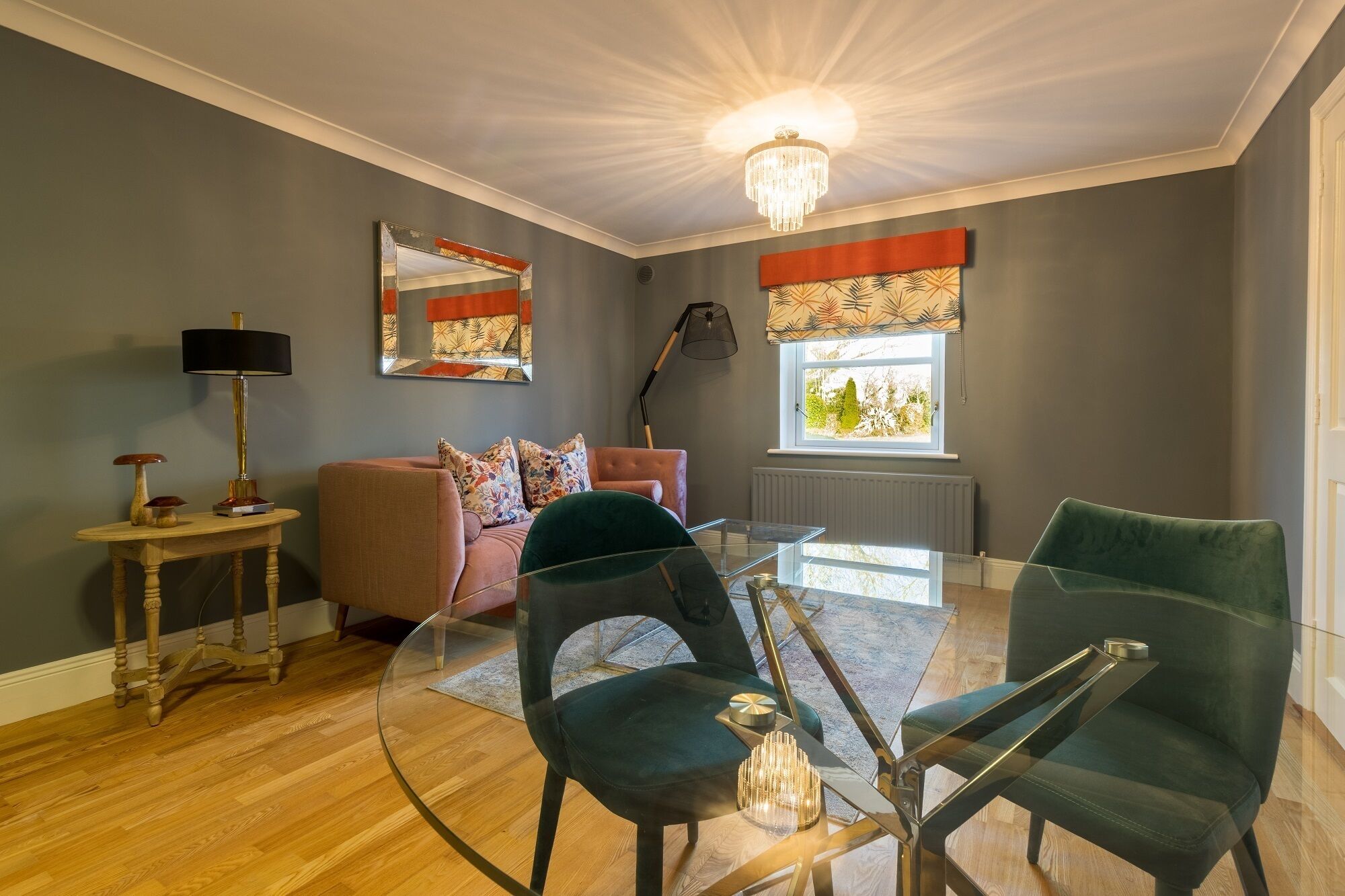 A modern living room with a glass coffee table, two green chairs, a pink sofa, and a chandelier.