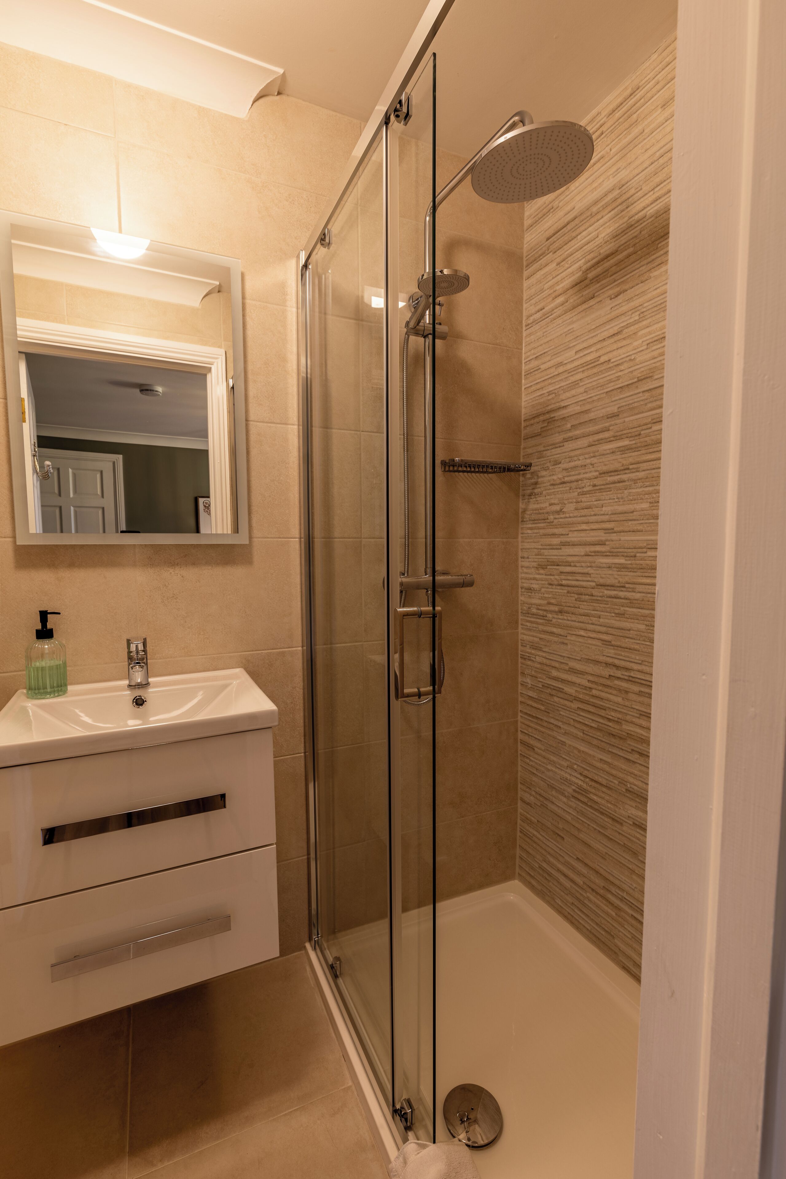 Un baño moderno con ducha de vidrio, un lavamanos blanco con dispensador de jabón y un espejo sobre el lavamanos.