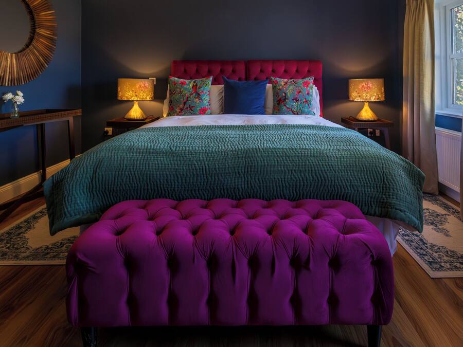 A bedroom with a tufted purple bench, a red headboard, and a teal bedspread.