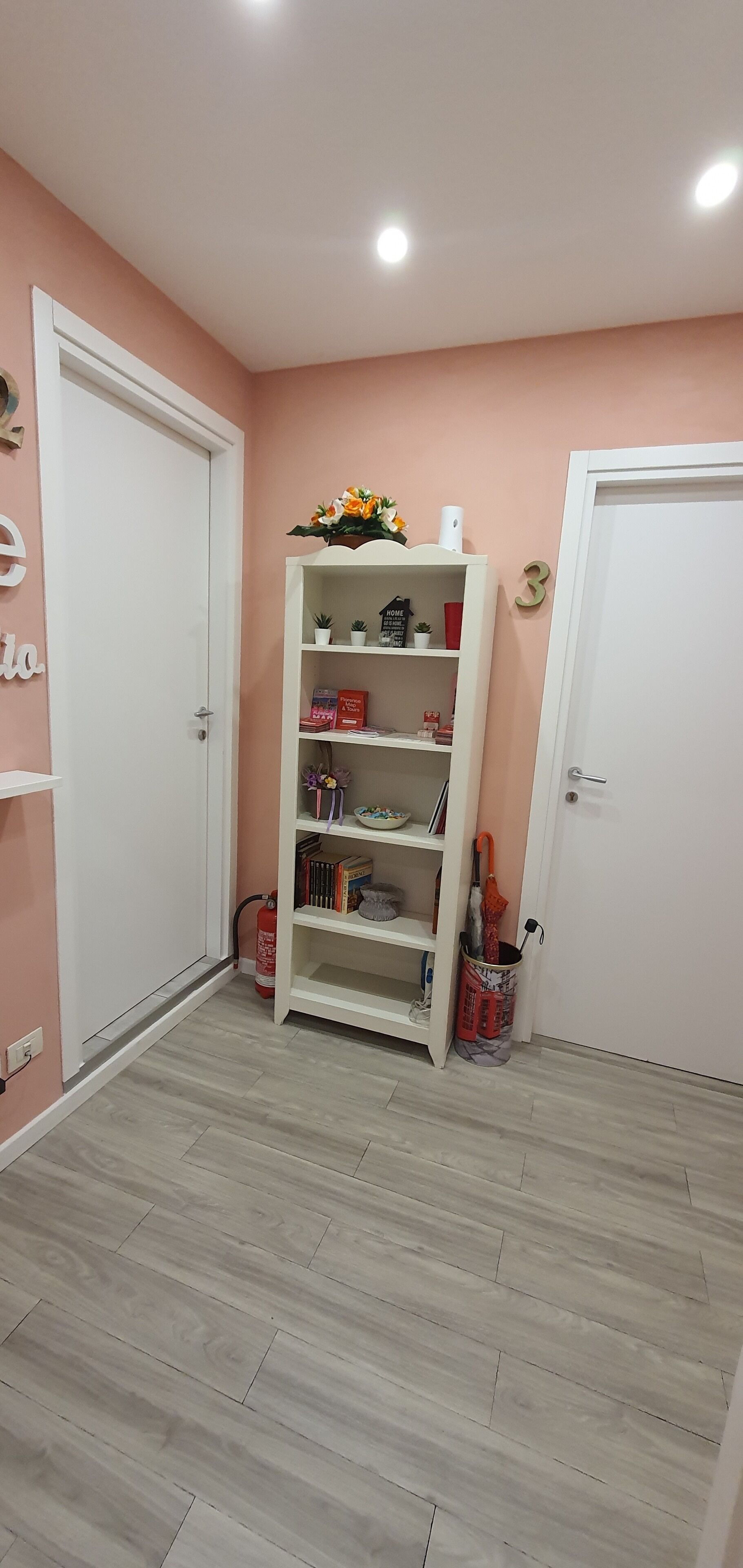 A hallway with a white door, a white shelf unit with various items, and a fire extinguisher on the floor.