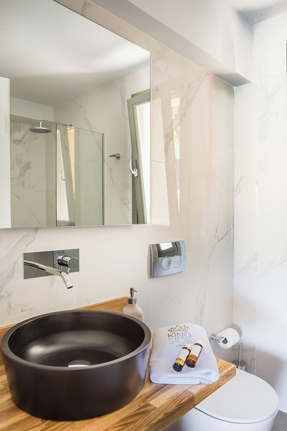 Une salle de bain moderne avec un mur en marbre, un lavabo noir, un plan de toilette en bois, des toilettes et un grand miroir.