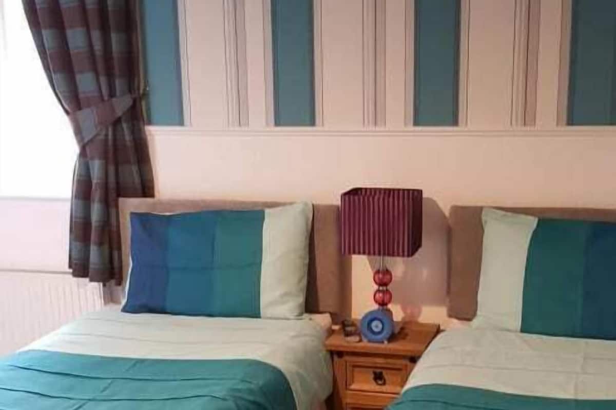 A bedroom with two beds, striped wallpaper, a framed picture on the wall, and a bedside table with a lamp.