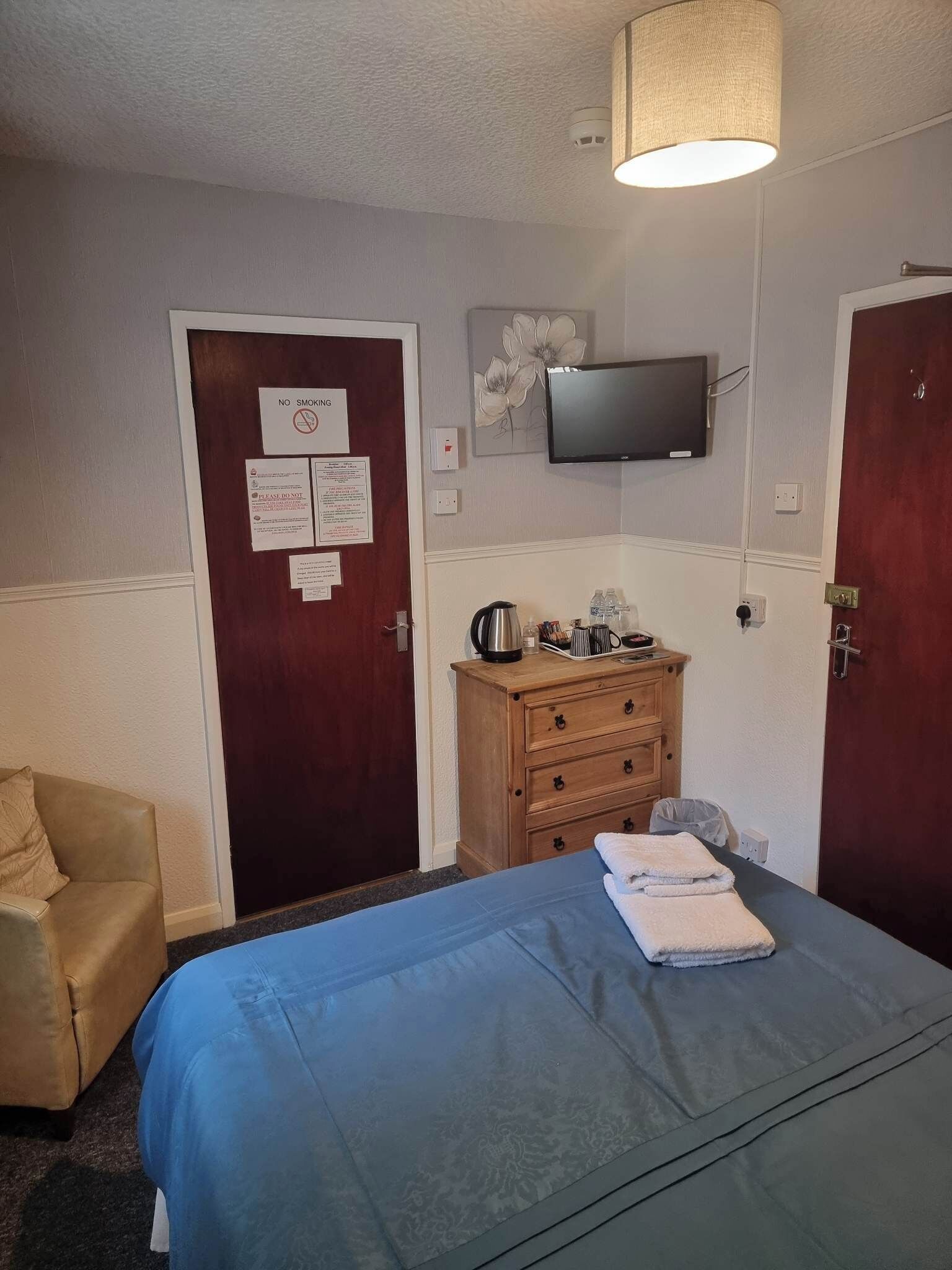 Una camera d'albergo con un letto, una cassettiera in legno, un televisore a parete e una porta con il cartello 'No Smoking'.