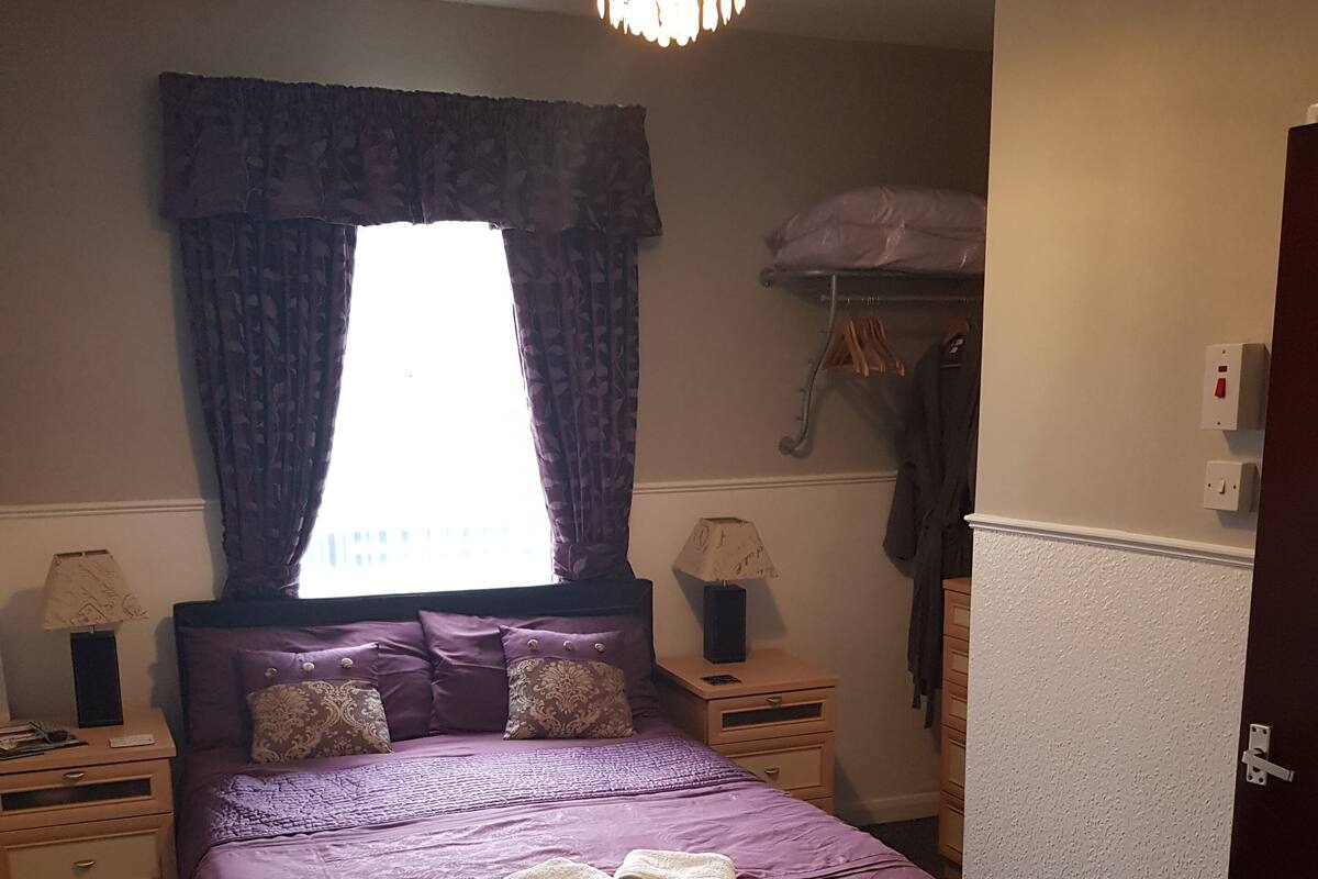 A bedroom with a bed, two bedside tables, a lamp, a window with curtains, a hanging coat rack, and a door.