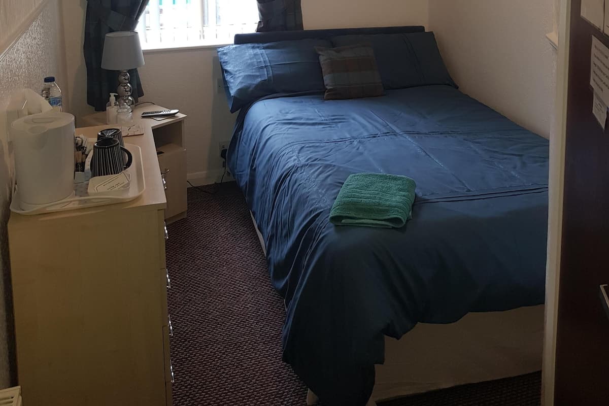 A compact hotel room with a single bed, a desk with a lamp, a hanging coat rack, and a window with curtains.