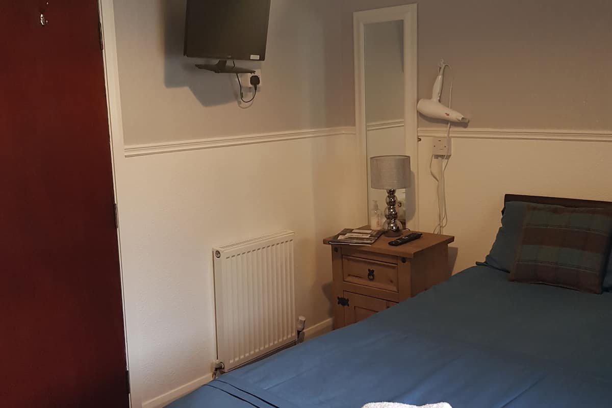 A bedroom with a bed, a TV mounted on the wall, a bedside table with a lamp, and a radiator.