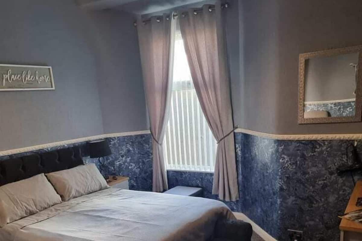 A bedroom with a bed, a bench, a window with curtains, a wall-mounted mirror, and a sign on the wall.