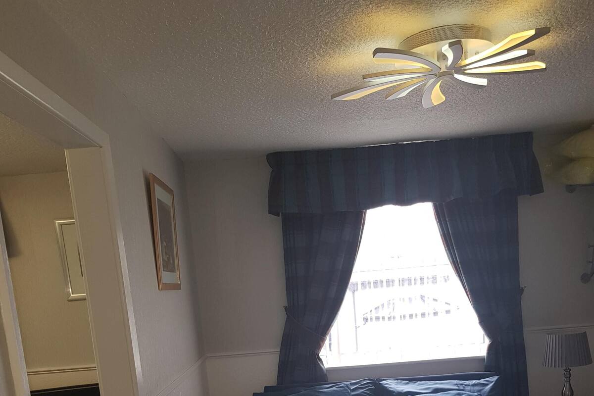 A bedroom with a bed, a window with curtains, a ceiling light fixture, and a nightstand with a lamp.