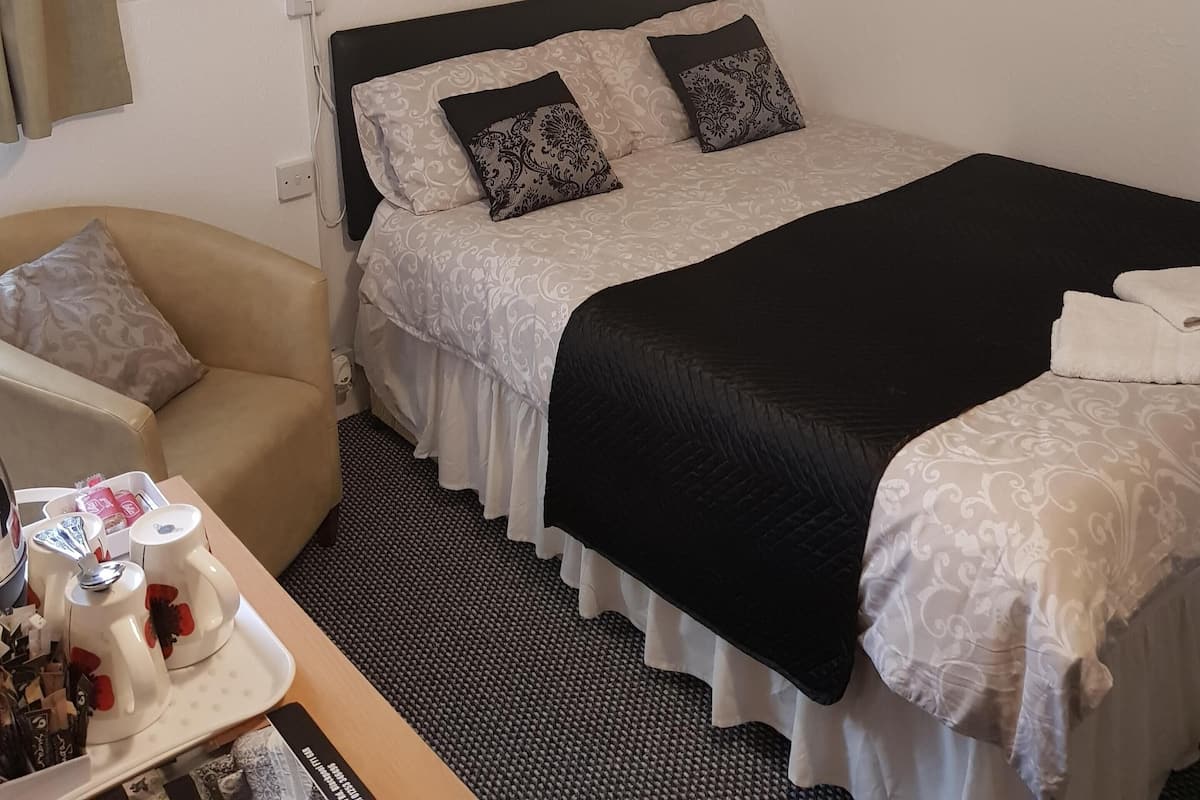 A hotel room with a bed, a bedside table with a tray of mugs and a cup, a chair, and a mirror on the wall.