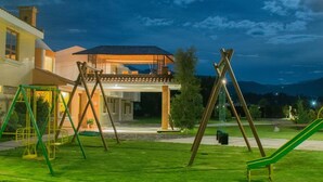 A playground with swings and a slide, a house with a balcony, and a mountain in the background. - Hacienda Grande Hotel (San Cristóbal Totonicapán)