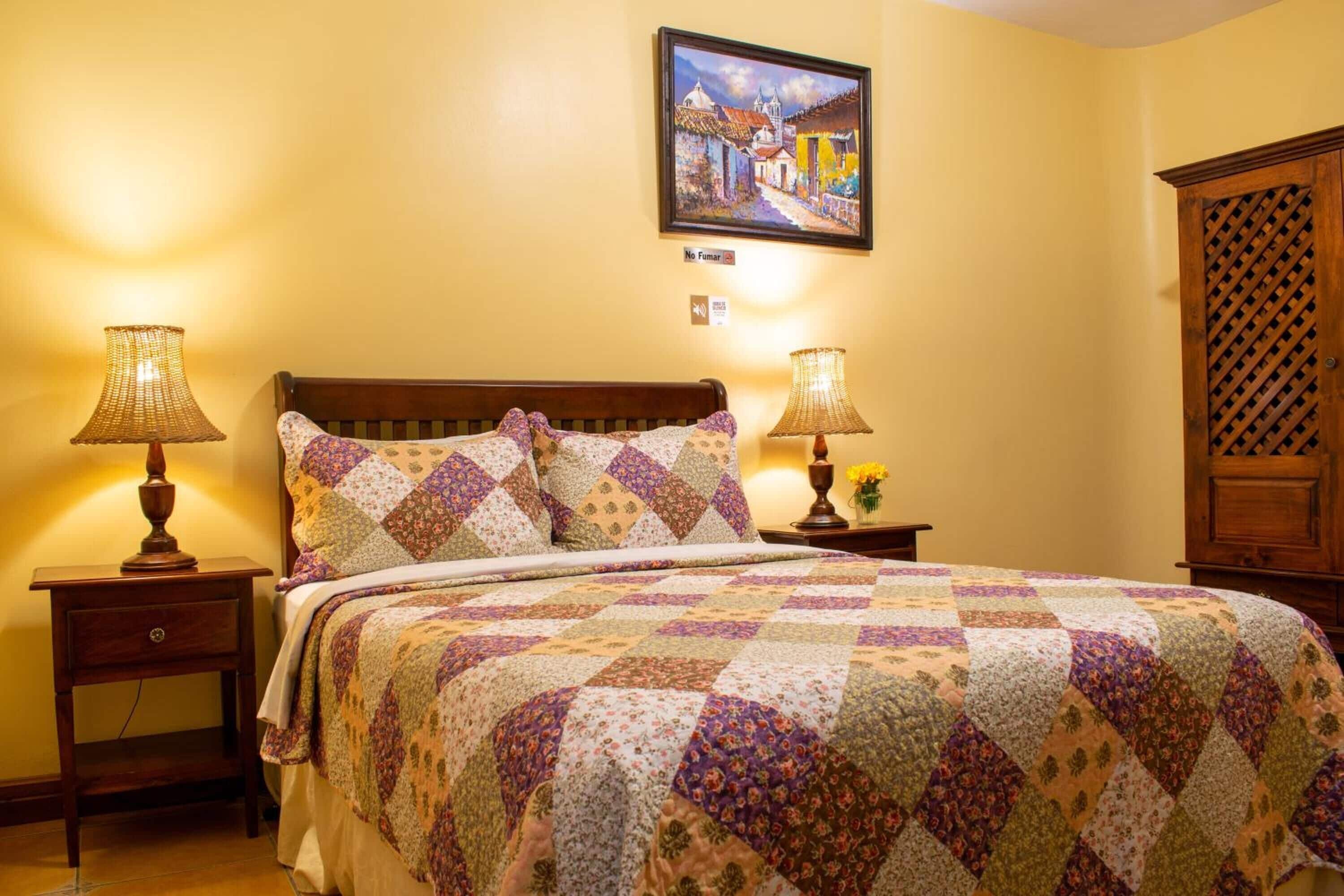 A neatly made bed with a quilt, two bedside lamps, a nightstand, a framed painting, and a wooden cabinet.