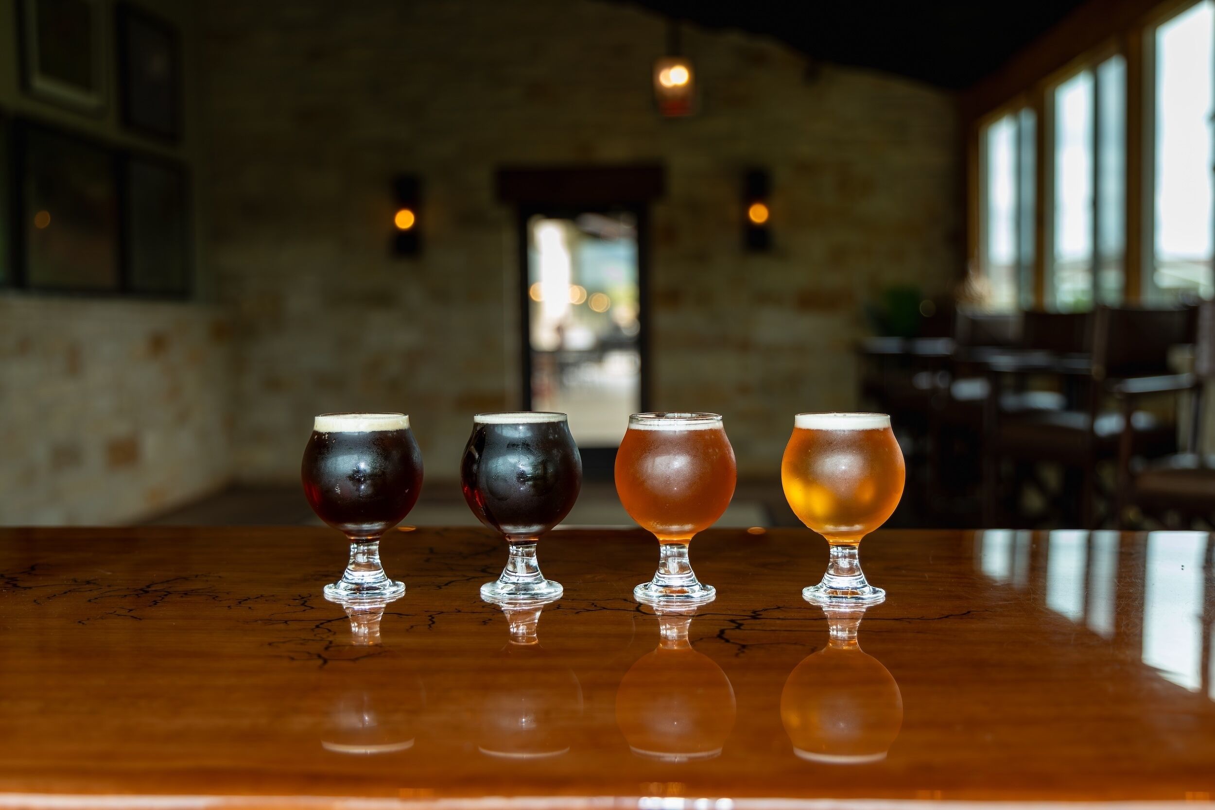 Four glasses of beer on a wooden bar counter in a dimly lit room with brick walls and large windows.