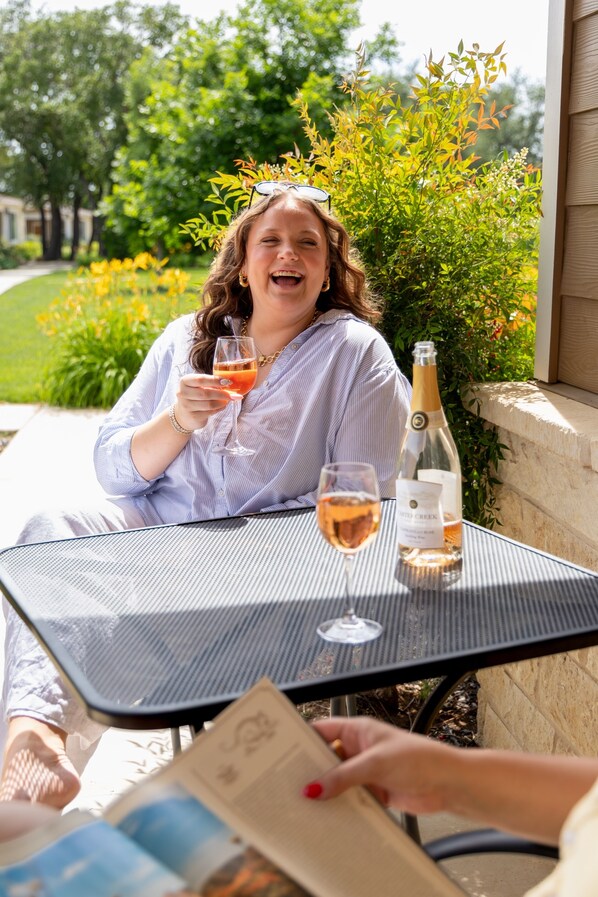 A person seated at an outdoor table with a bottle and glasses, suggesting a dining or social setting.