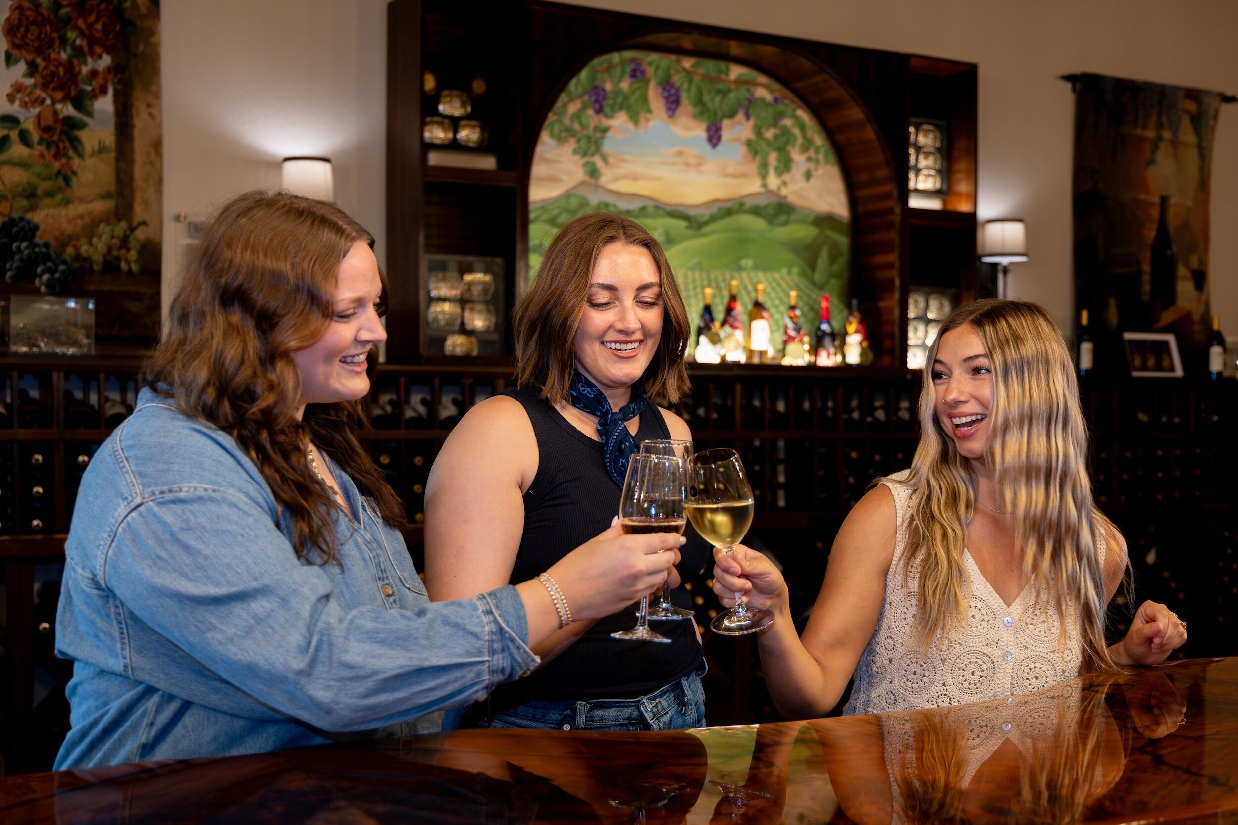 Three people toasting with wine glasses at a bar with a mural and wine bottles in the background.