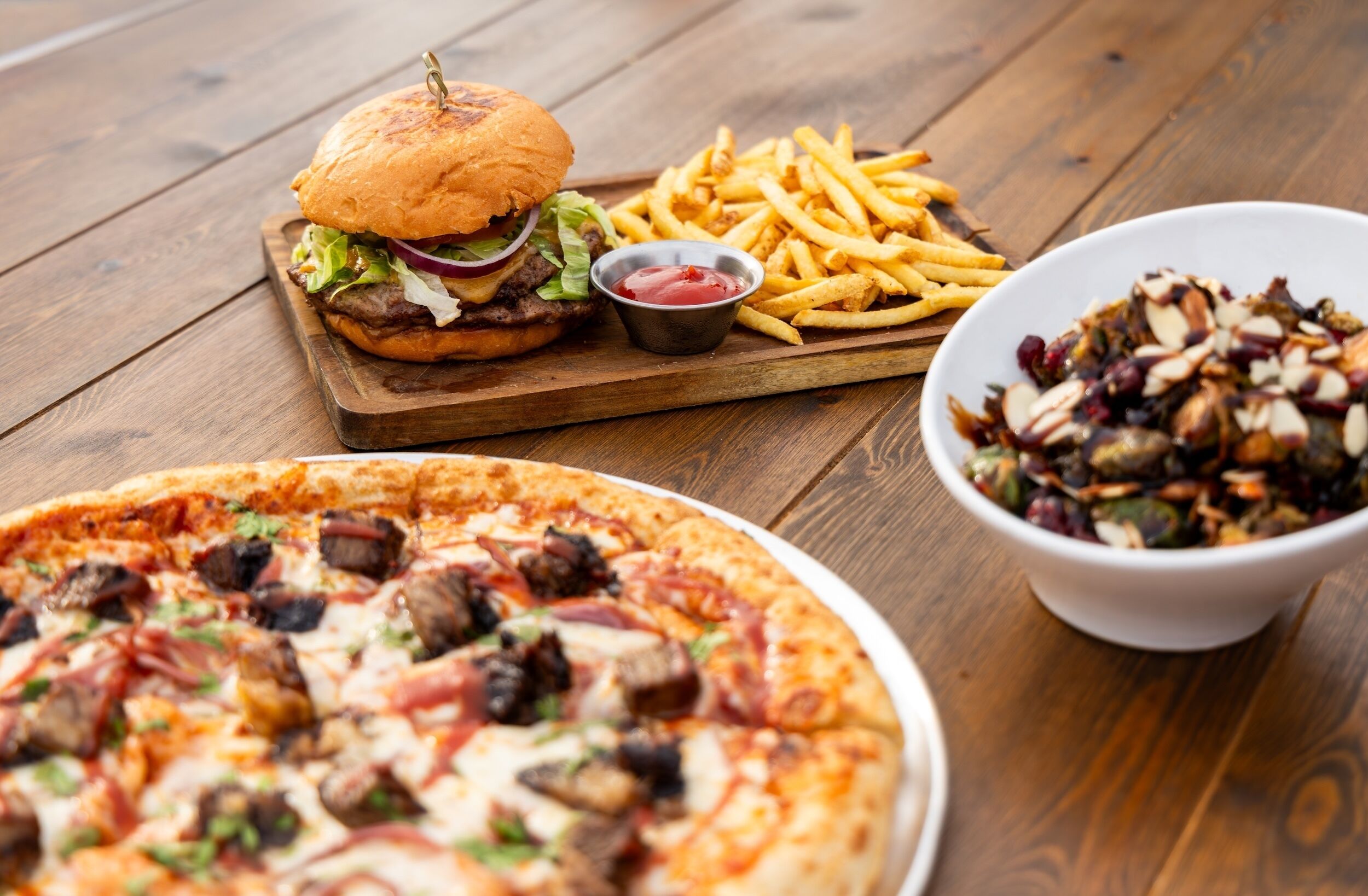 A wooden table with a burger, fries, a bowl of salad, and a pizza.