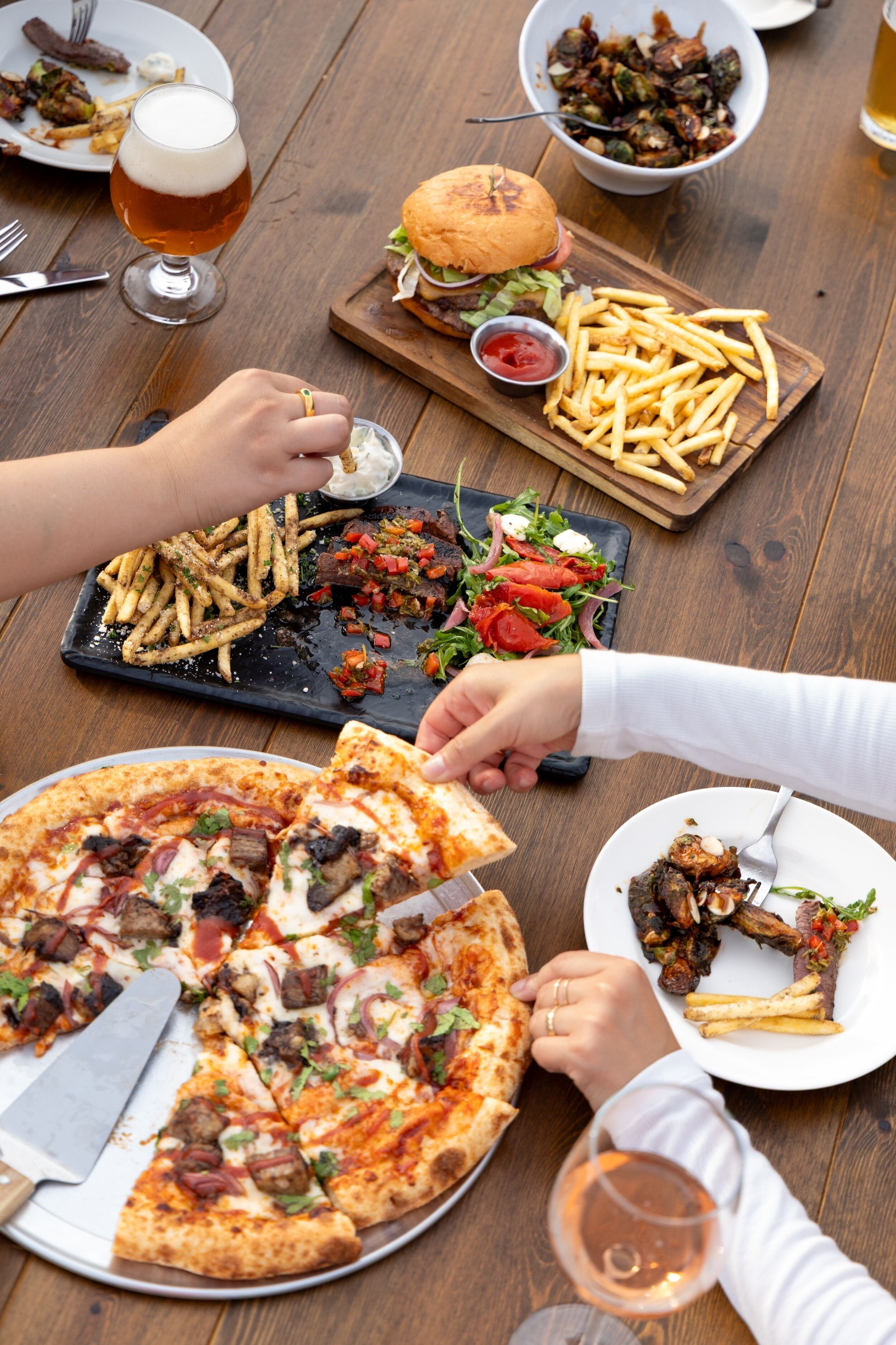 Uma mesa de jantar com pizza, hambúrguer, batatas fritas, salada e um copo de cerveja.
