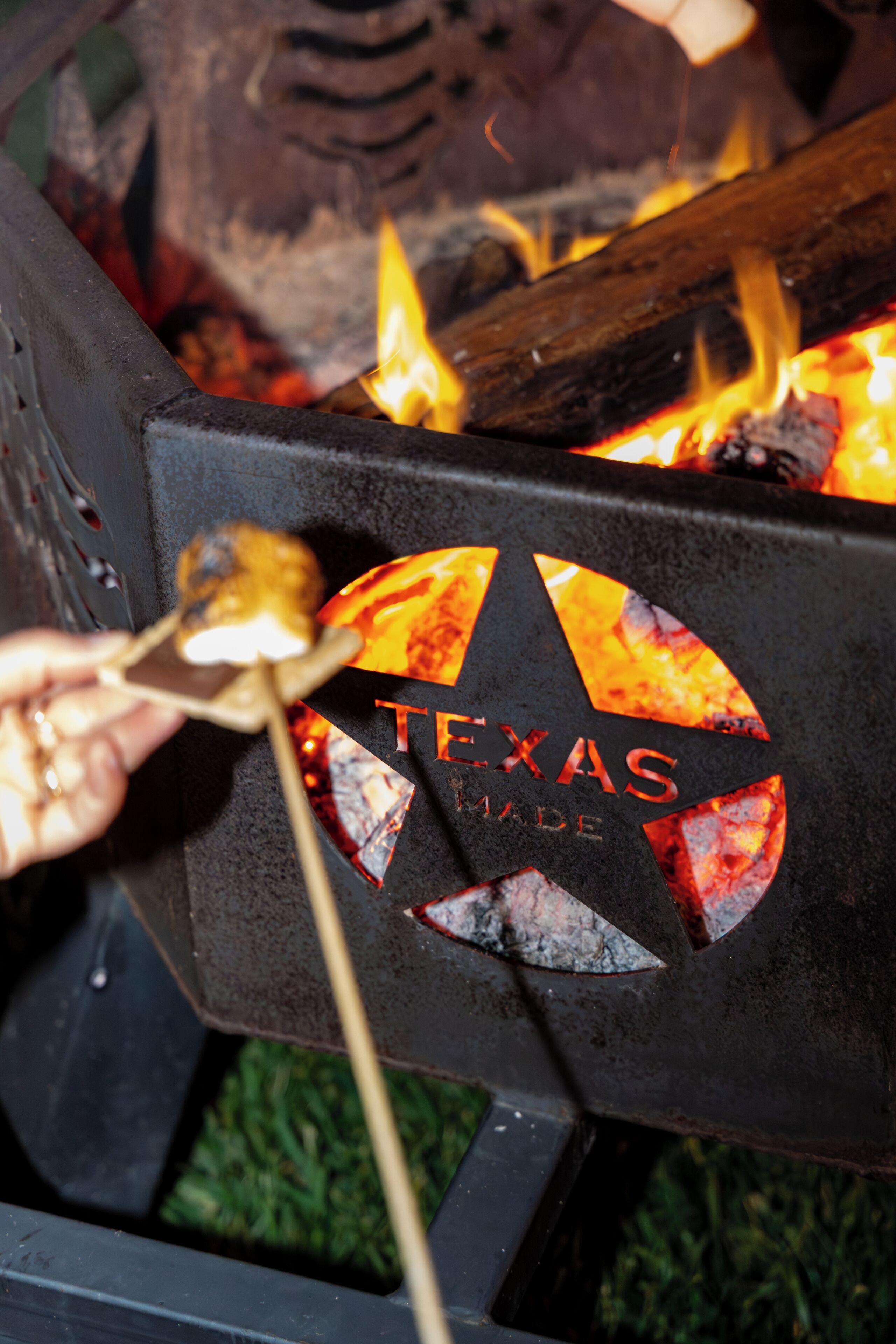 Una parrilla con la marca 'TEXAS MADE', una mano sosteniendo un pincho con comida y llamas visibles.