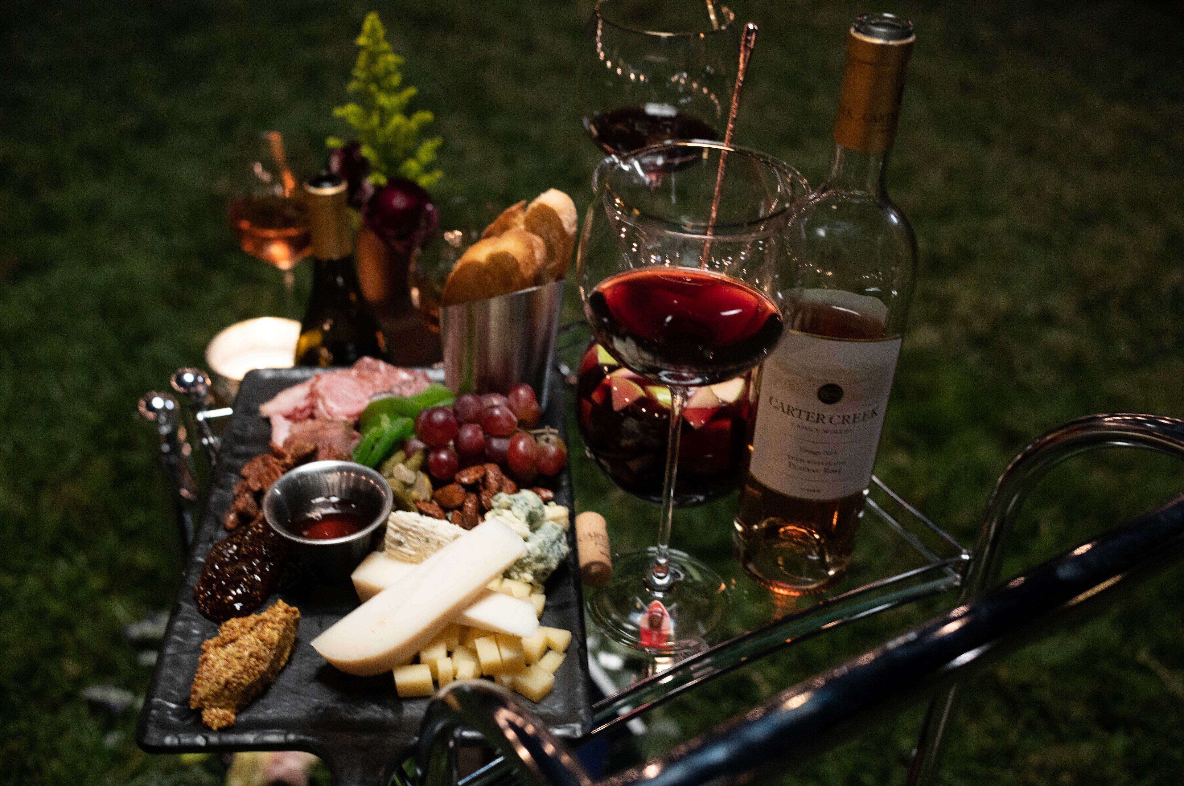A picnic setup with a variety of cheeses, meats, and fruits on a tray, accompanied by wine glasses and a bottle of wine.
