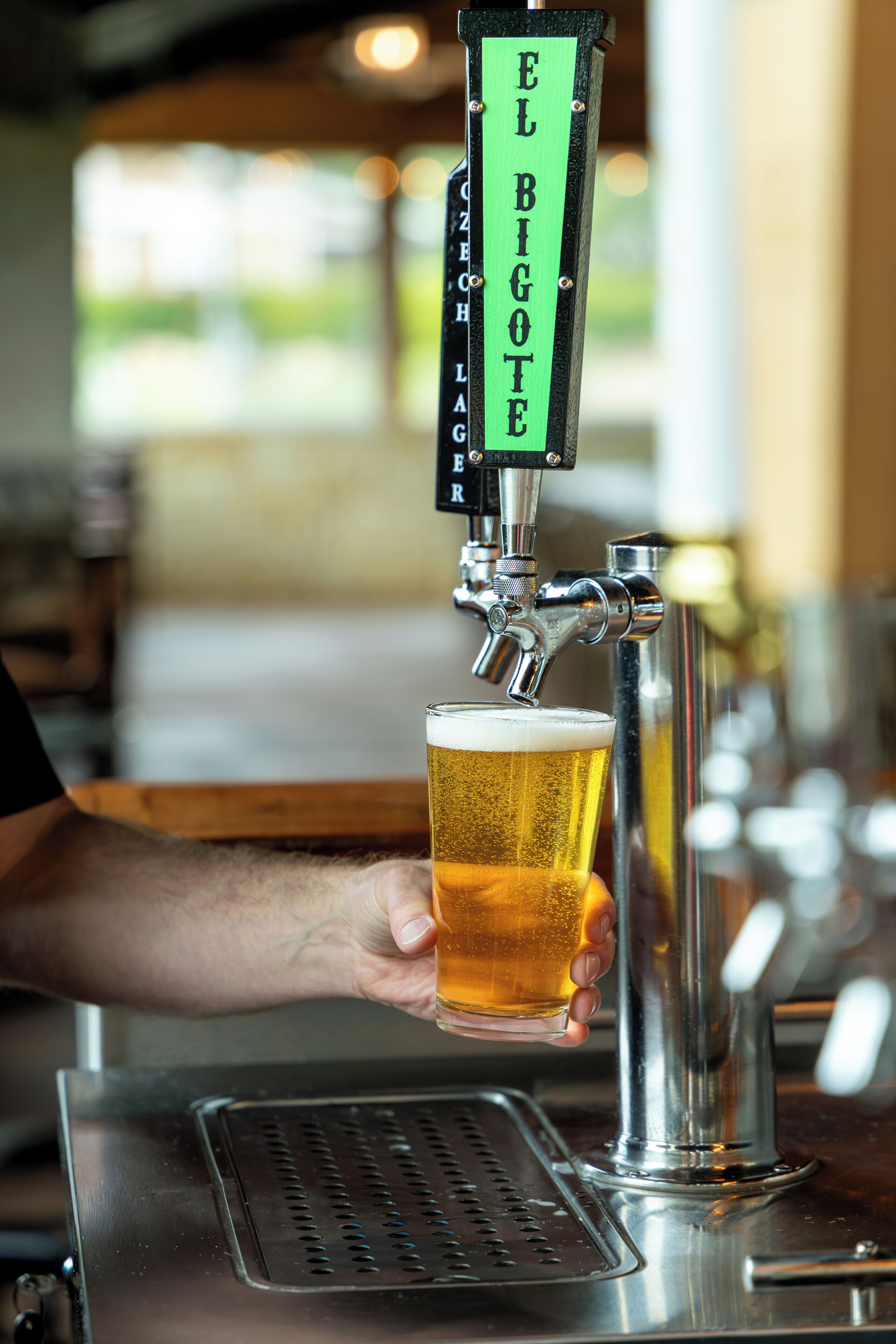 A hand holding a glass of beer at a tap labeled 'EL BIGOTE.'