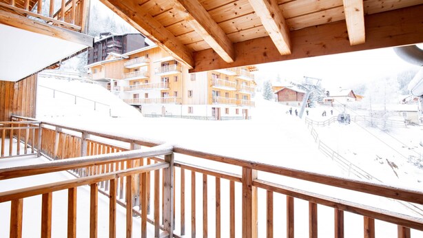Ein Holzbalkon mit Metallgeländer, der einen verschneiten Ort mit mehrstöckigen Gebäuden überblickt.