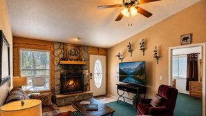A cozy living room with a fireplace, a television, a ceiling fan, and a stone wall. - Hilltop Cabin (Ruidoso)