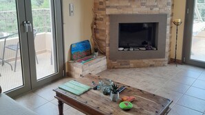 Una sala de estar con chimenea de piedra, televisor montado arriba y balcón visible a través de puertas corredizas de vidrio.