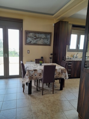 A dining area with a table set for four, a painting on the wall, and a kitchen in the background. - Villa Kastania (Apokoronas)