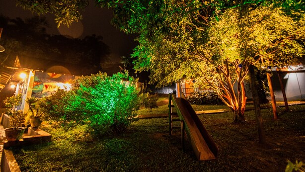 Un espacio al aire libre de noche con un banco de madera, un pequeño árbol y un edificio con ventanas iluminadas.
