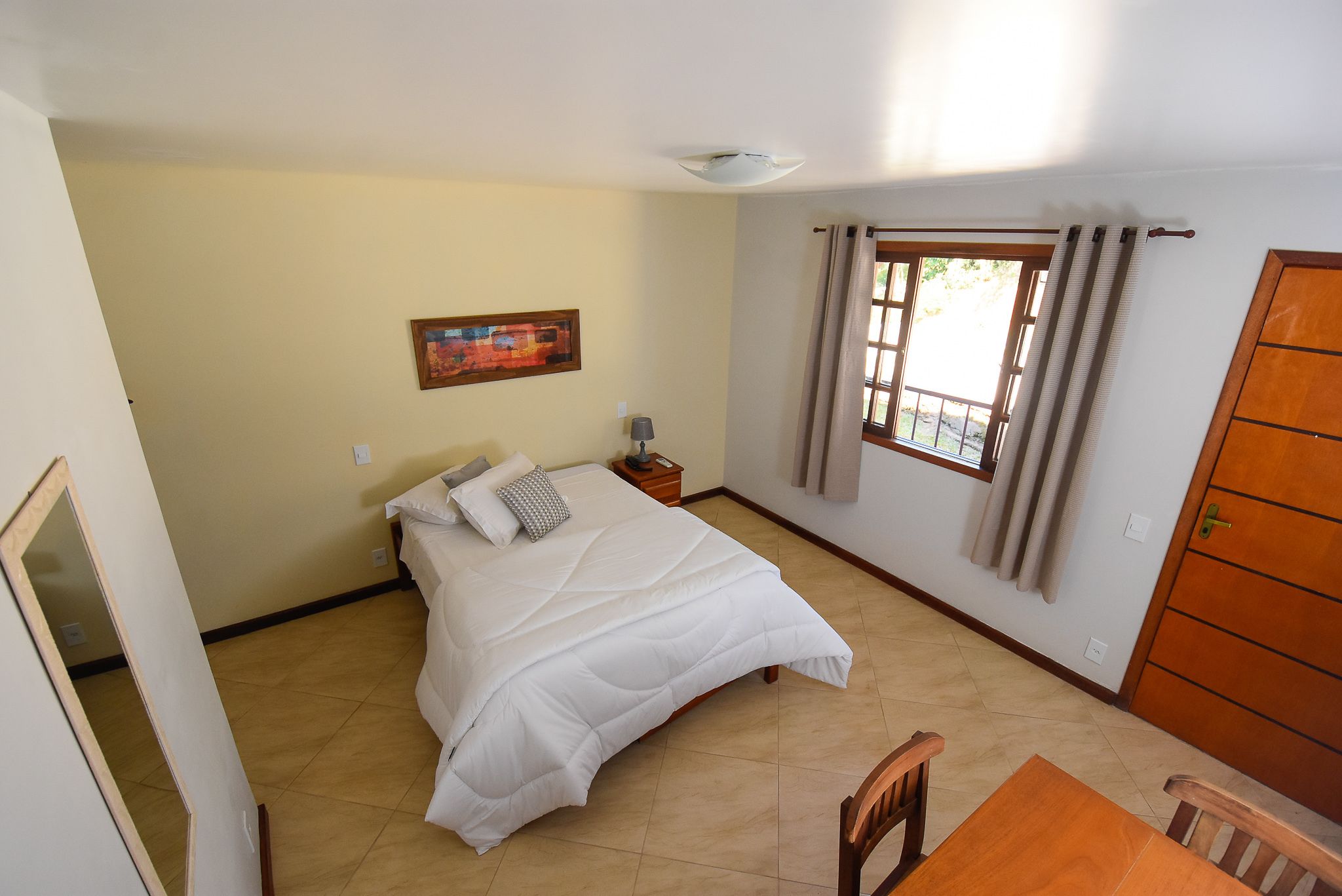 A bedroom with a bed, a nightstand, a lamp, a painting on the wall, a window with curtains, and a wooden door.