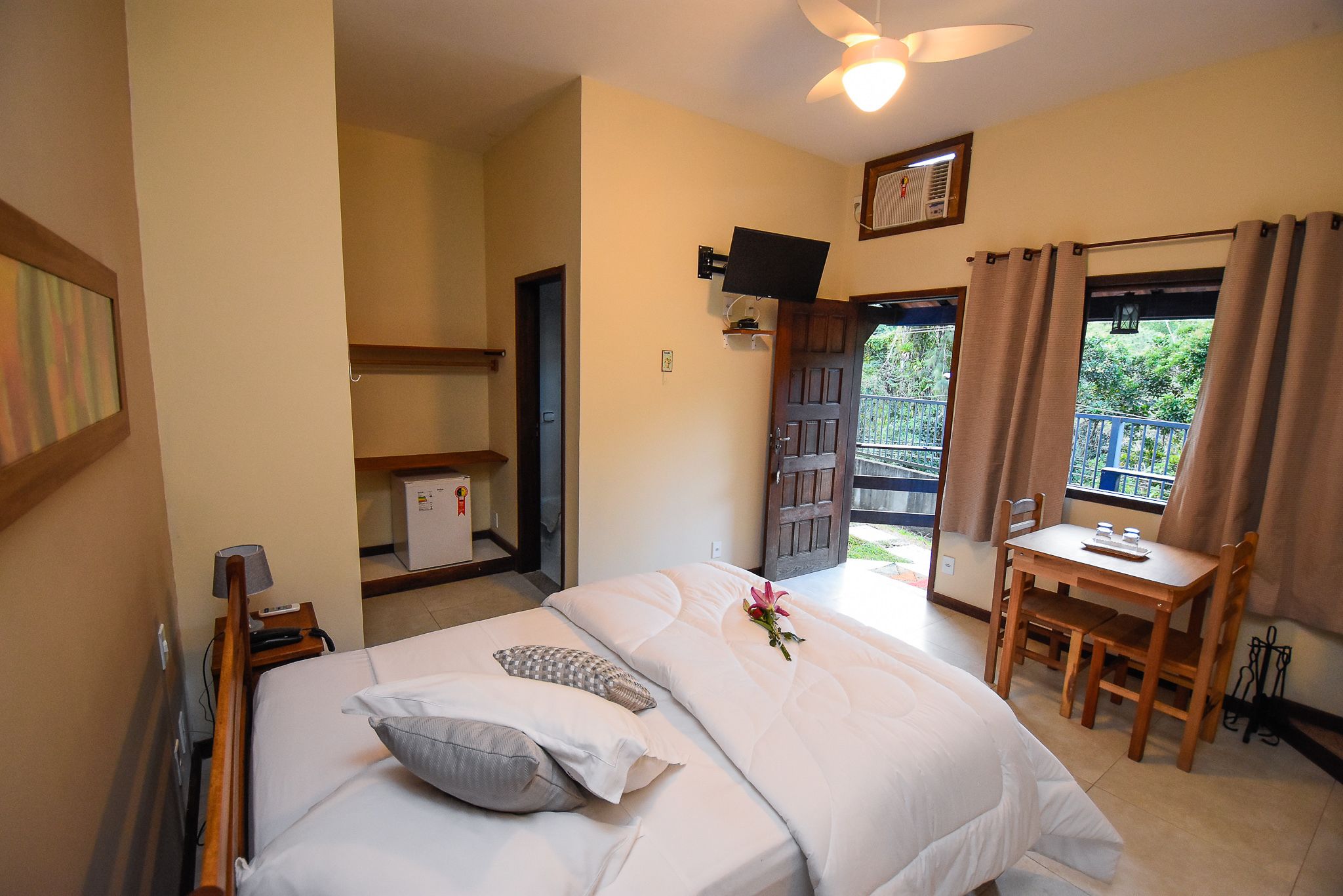 Un dormitorio con cama, mesita de noche, lámpara y un escritorio pequeño con una silla. Hay una ventana con cortinas y vistas al exterior con vegetación.