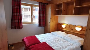 A wooden room with a bed, two red blankets, a window with plaid curtains, and a ceiling light.