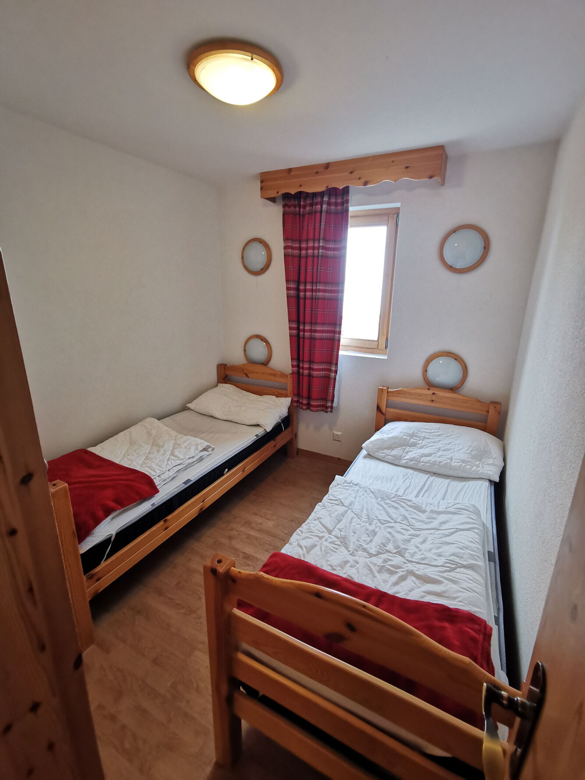 Une petite pièce chaleureuse avec deux lits superposés en bois, une fenêtre ornée d’un rideau rouge et un luminaire au plafond.