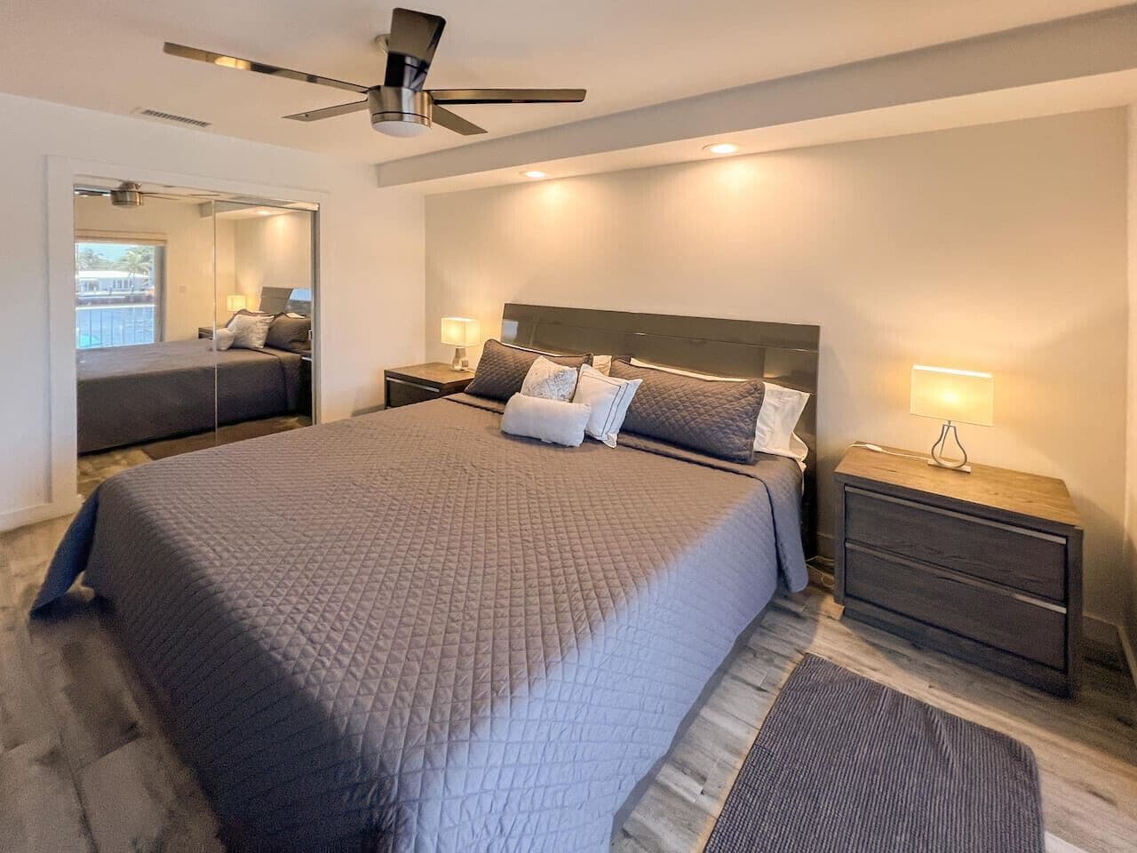 Une chambre moderne avec un grand lit, des tables de chevet, un ventilateur de plafond et une vue sur l’extérieur, visible à travers une porte vitrée.
