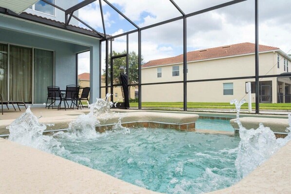 A swimming pool with water jets, a covered patio with chairs, and a view of houses in the background. - 8950 Mermaid & Batman Rooms, Game Room! Pool (Kissimmee)