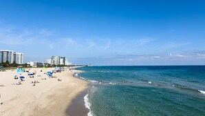 Una playa con cielos azules despejados, orilla arenosa y varios edificios al fondo.