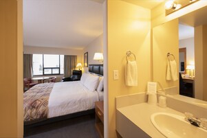 Quarto de hotel com cama, mesa de cabeceira, abajur e banheiro com pia e espelho.
