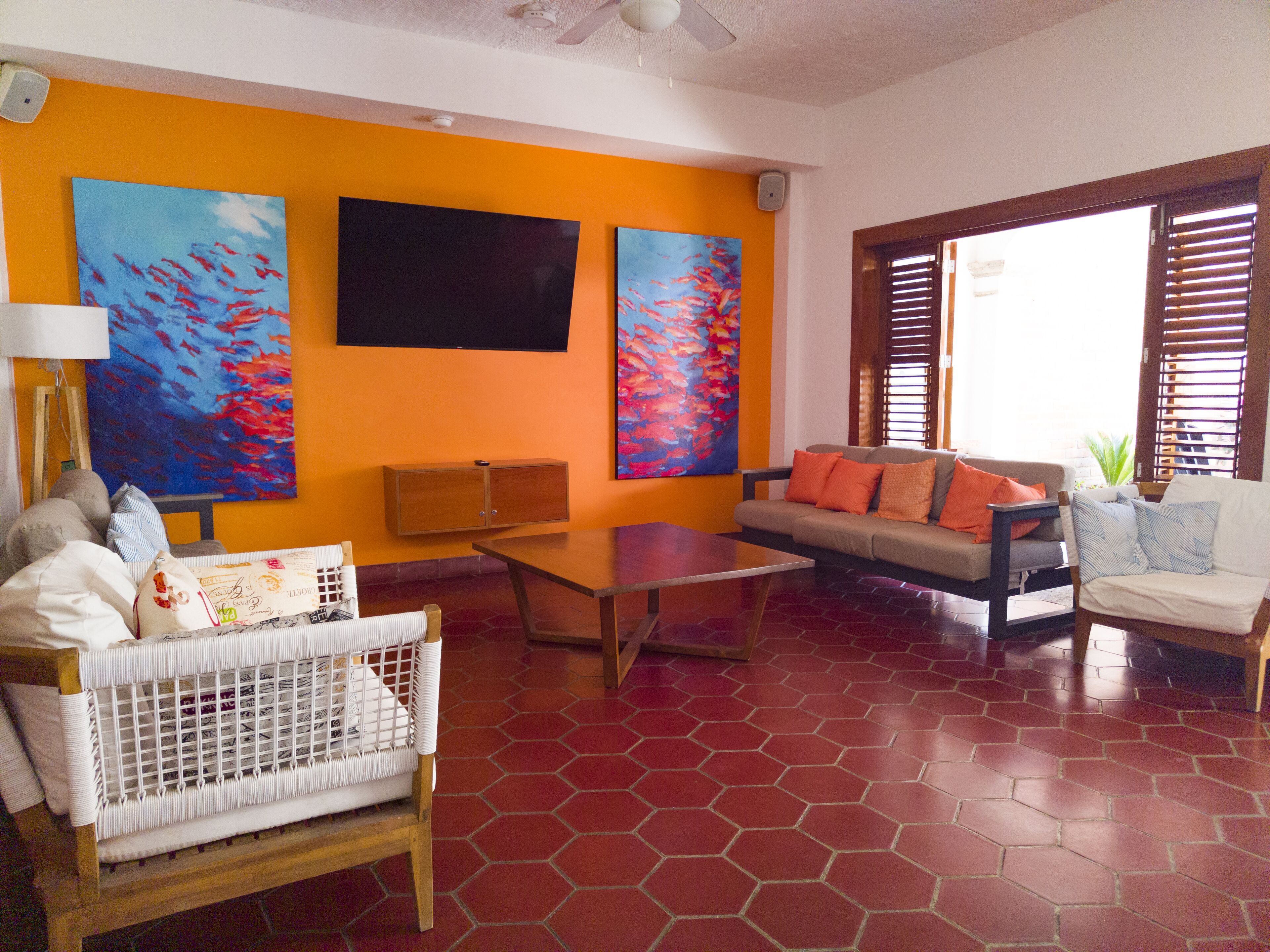 Una sala de estar con un sofá, una mesa de centro y un sillón. Hay un televisor montado en la pared, y dos cuadros encima.