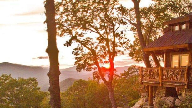 Una cabana de fusta amb un balcó, envoltada d'arbres i muntanyes, durant el capvespre.